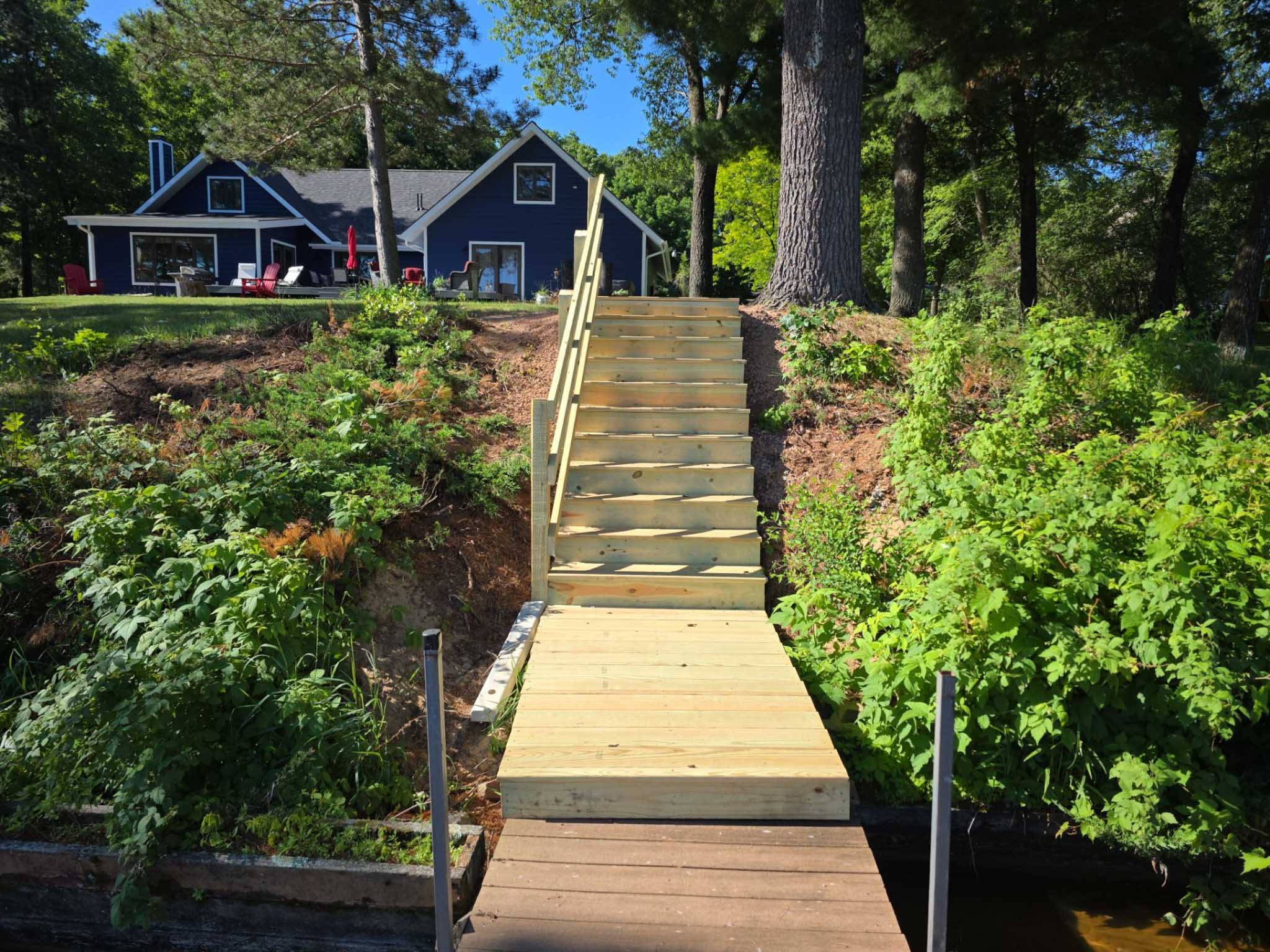 Wooden stairs connect a lakeside dock to a blue house—ask your Composite Deck Builder Wisconsin.
