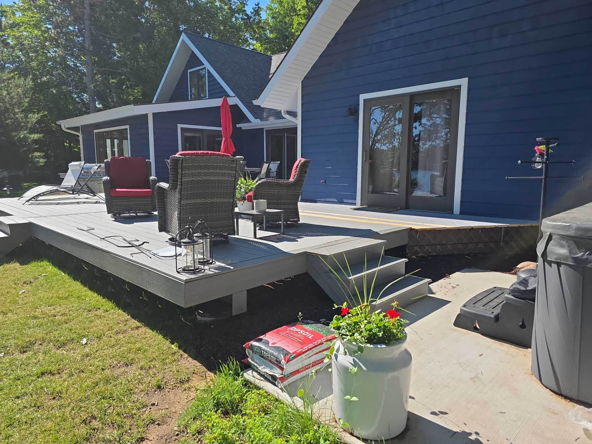 Patio with chairs, umbrella, grill, and soil bags by a blue house from a Composite Deck Builder.