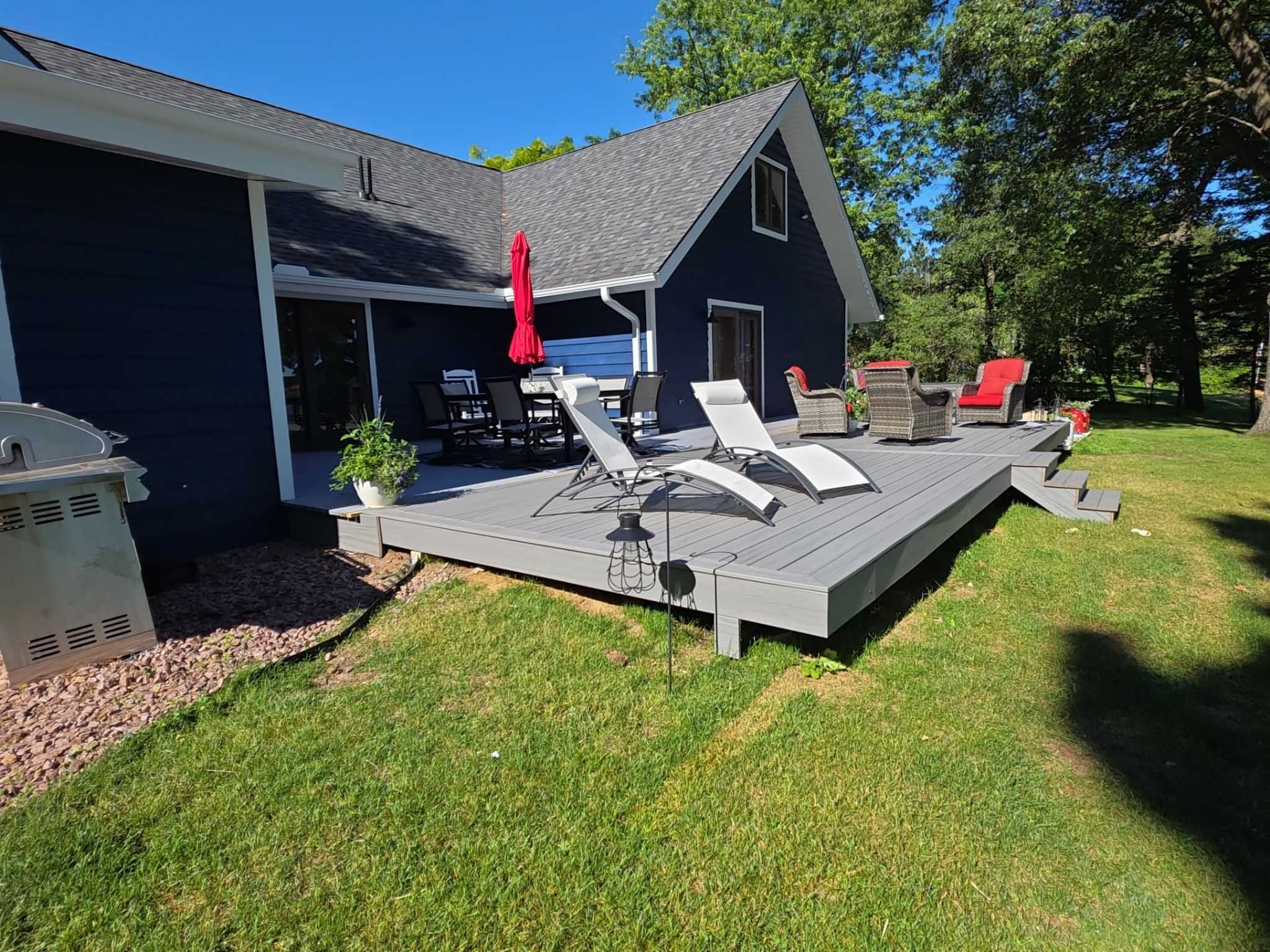 Gray patio deck by a dark blue house, built by Composite Deck Builder Wisconsin, with red umbrella.