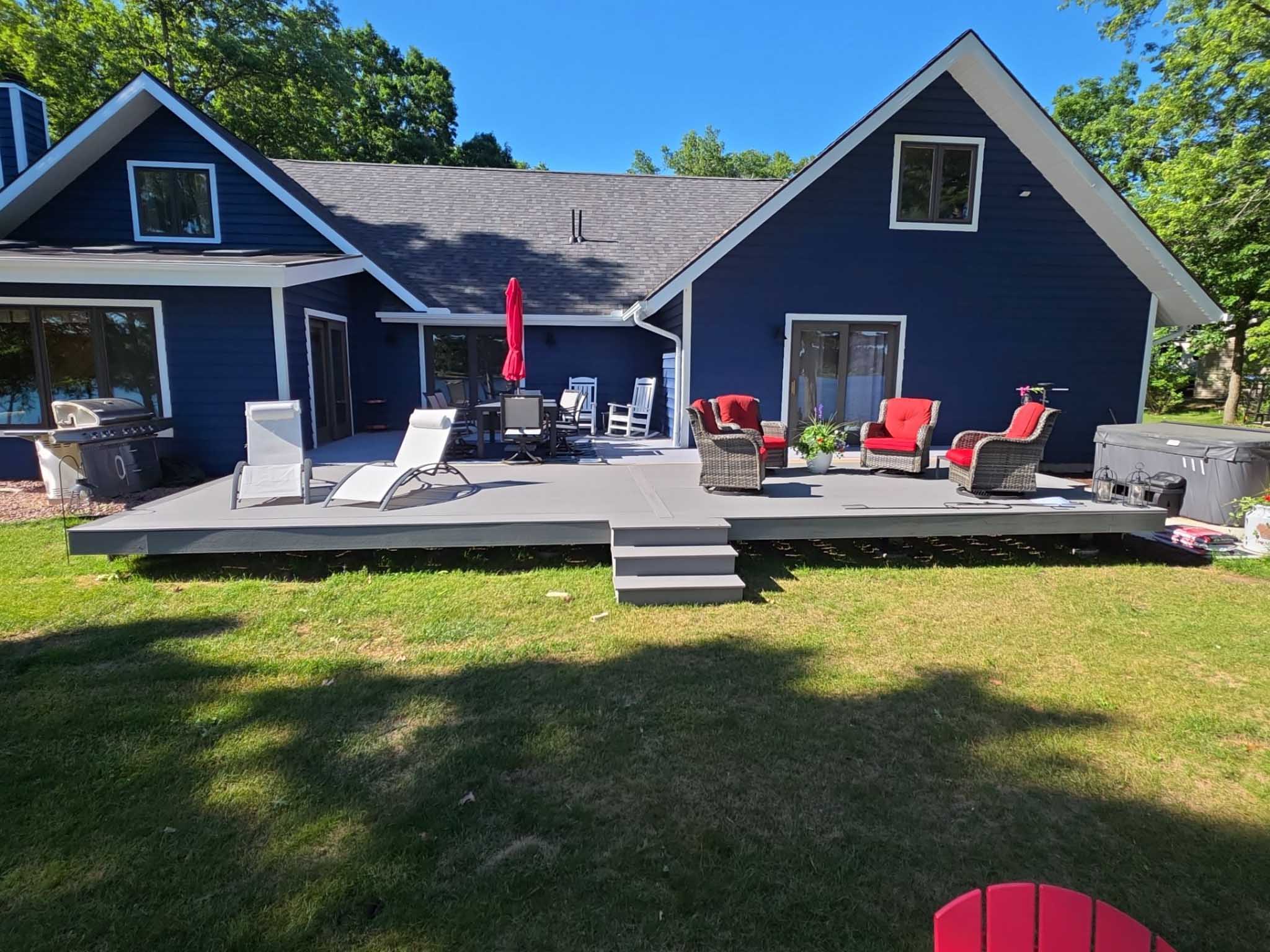Dark blue house with a composite deck, red chairs, lounge chairs, and a red umbrella in sunlight.