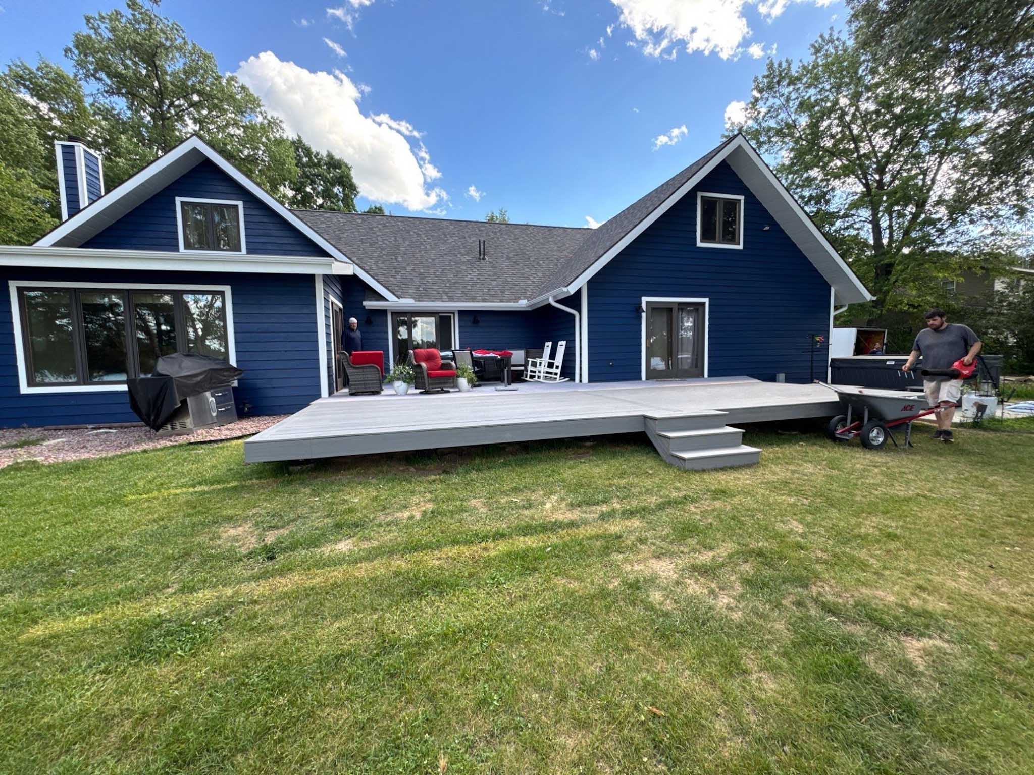 Blue house with gray composite deck, patio furniture, and a man walking near grill and wheelbarrow.