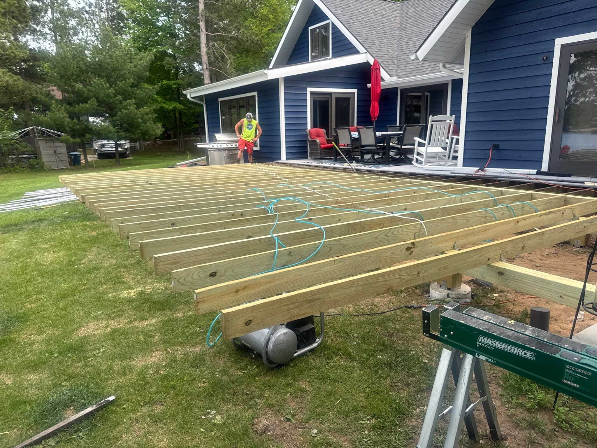 A Composite Deck Builder Wisconsin crafts a sturdy wooden deck frame by a blue backyard house.