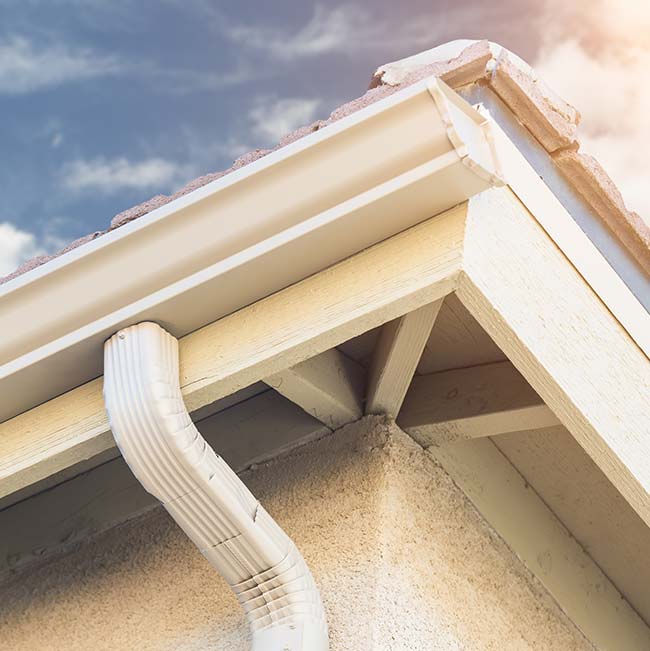 Close-up of Wisconsin house corner with roofing, gutters, and downspout against a blue sky.