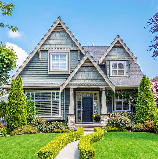 A green house with gabled roofing, manicured lawn, and trimmed hedges under a bright blue sky.