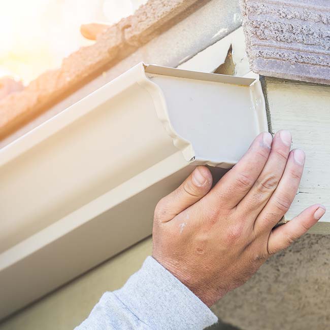 Hand installing white rain gutter on house roof edge in sunlight; ideal for roofing.