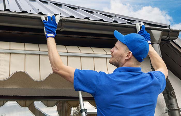 Man in blue uniform cleaning gutters on a house roof, ensuring roofing stays in top condition.