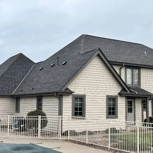 A two-story beige house with dark roof, white fence, and cloudy sky in the background.
