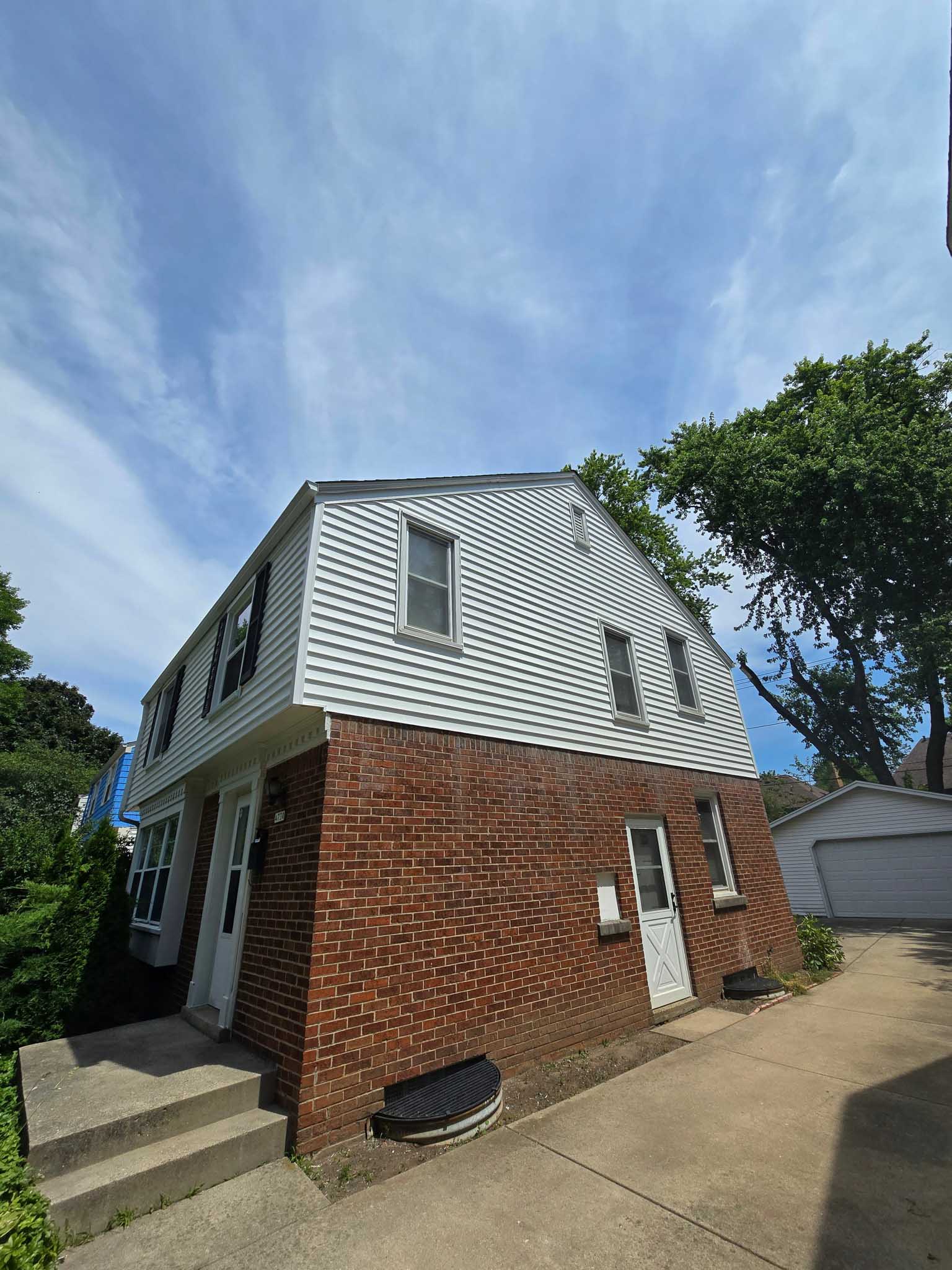 Two-story house: brick bottom, white siding by Siding Contractor Wisconsin, driveway, trees behind.
