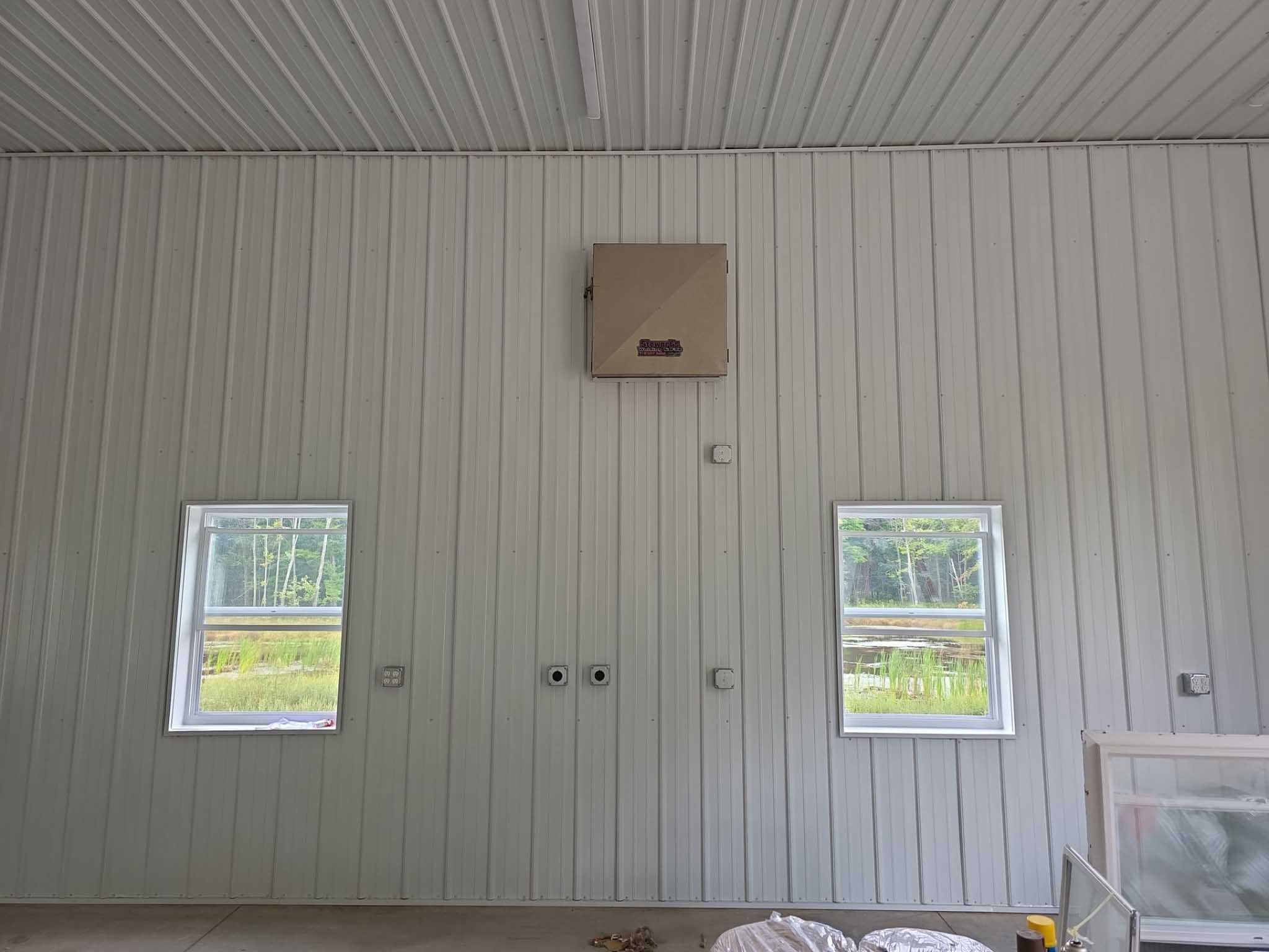 White paneled wall with two Wisconsin replacement windows, a brown box above, and greenery outside.