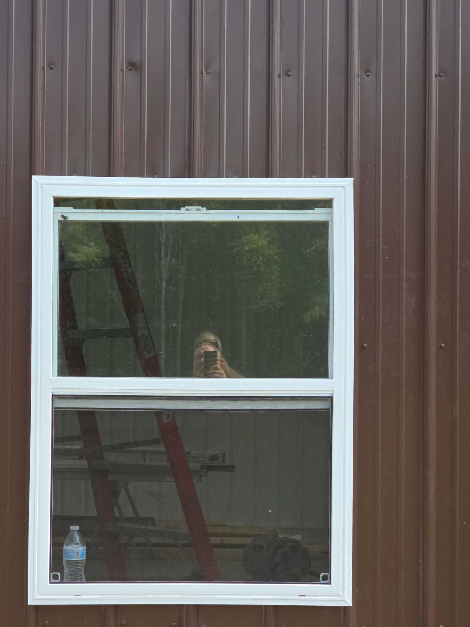 Person taking photo reflected in window on brown wall, ladder, bottle, Replacement Windows WI.