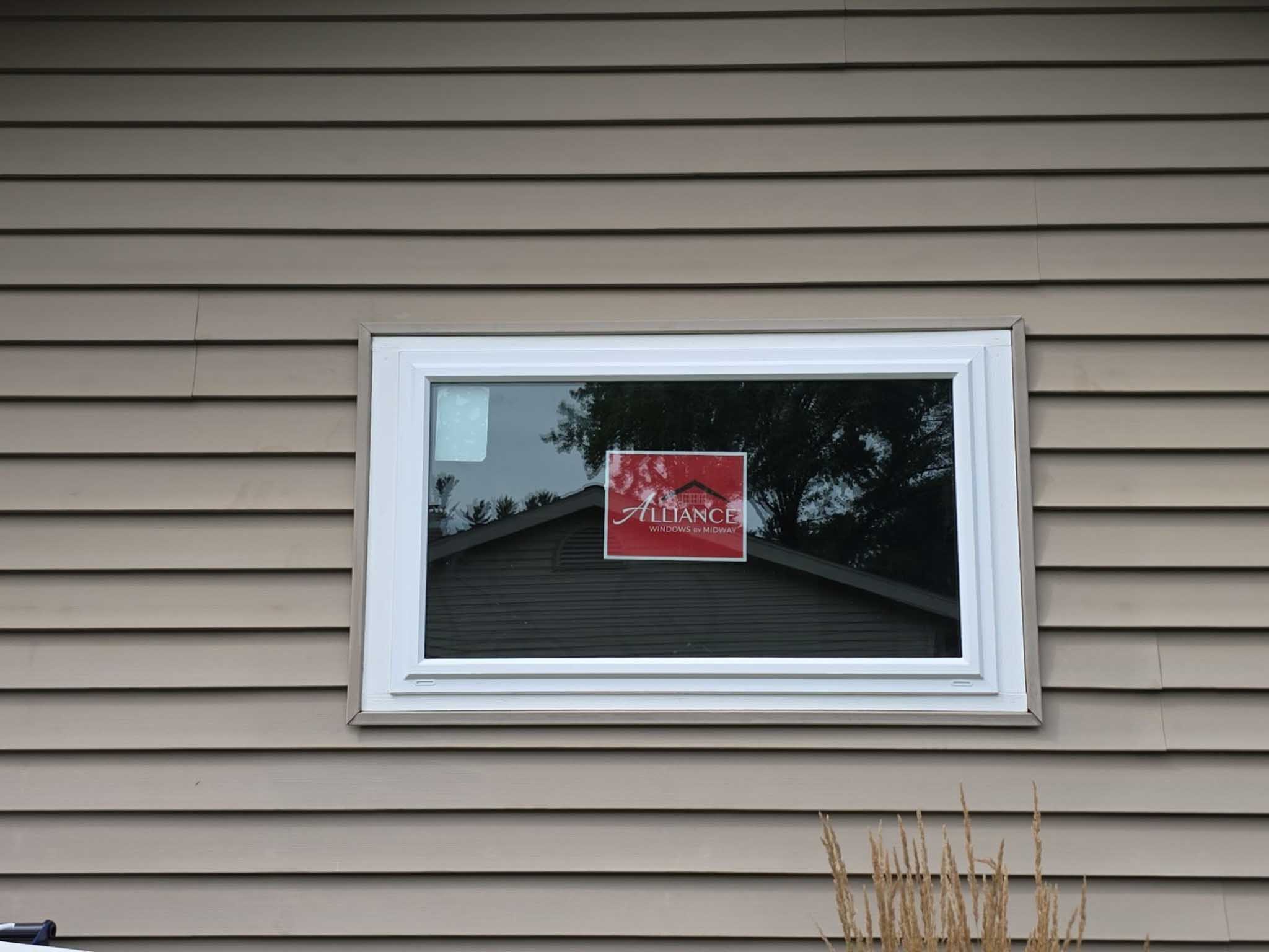 Rectangular window with red Alliance sign reflects house, trees—Replacement Windows Wisconsin.