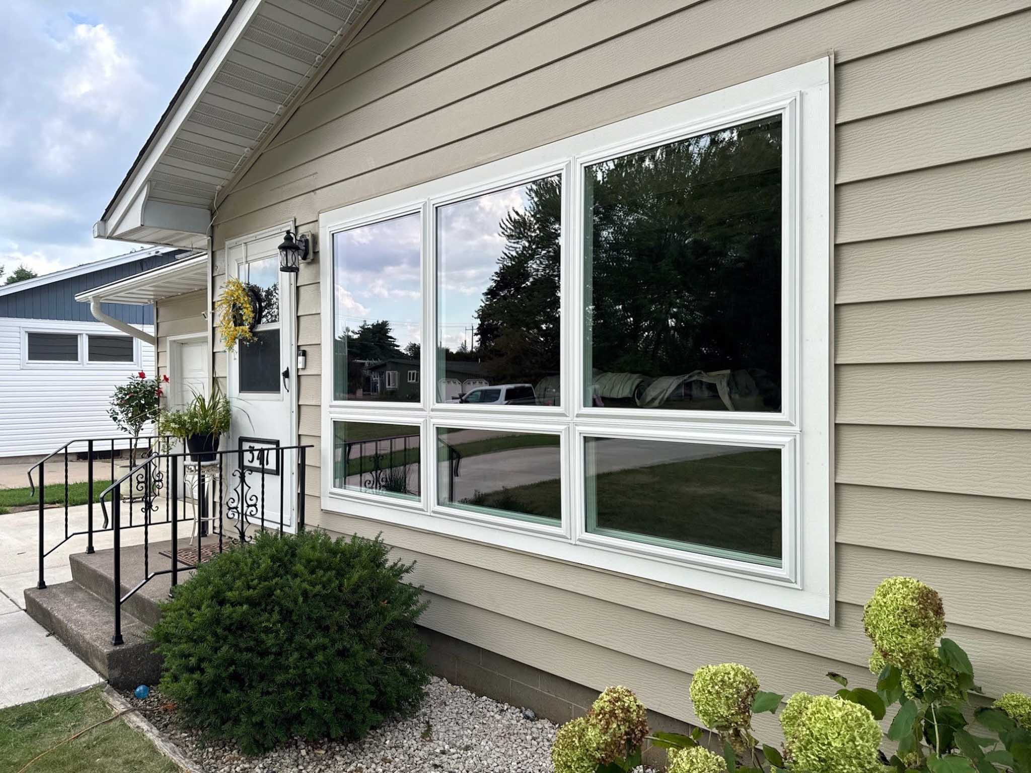 Beige house with white-framed replacement windows, a front porch, greenery, and cloudy sky.