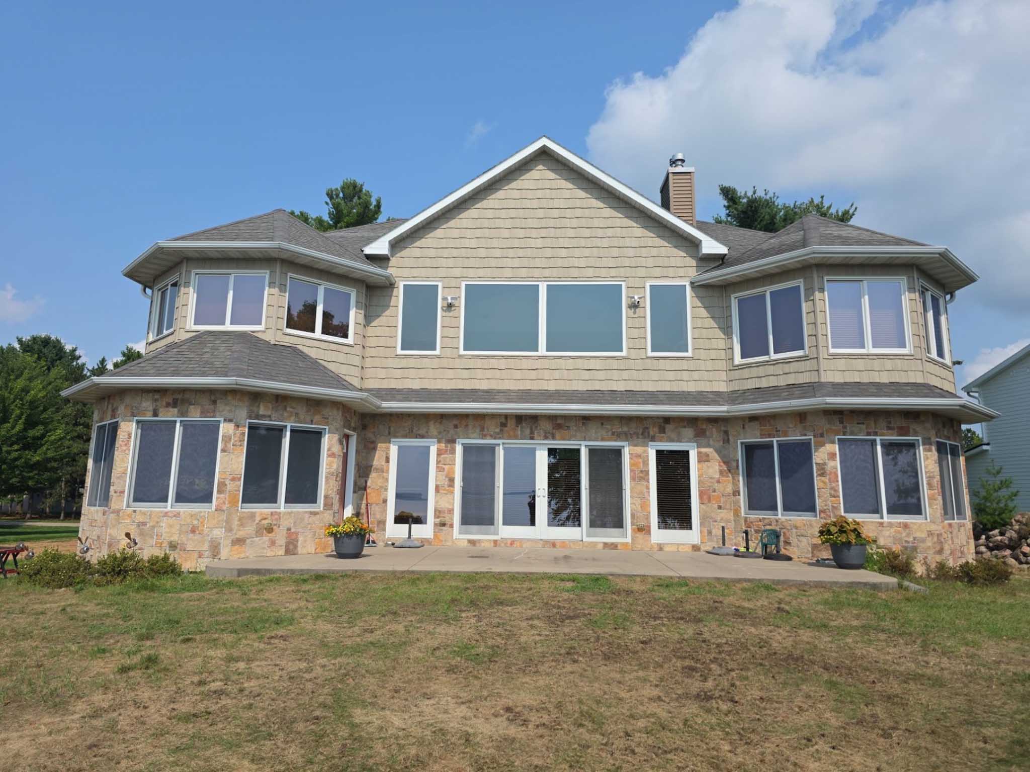 Two-story house with beige siding, stone facade, and Replacement Windows Wisconsin, dry grass lawn.