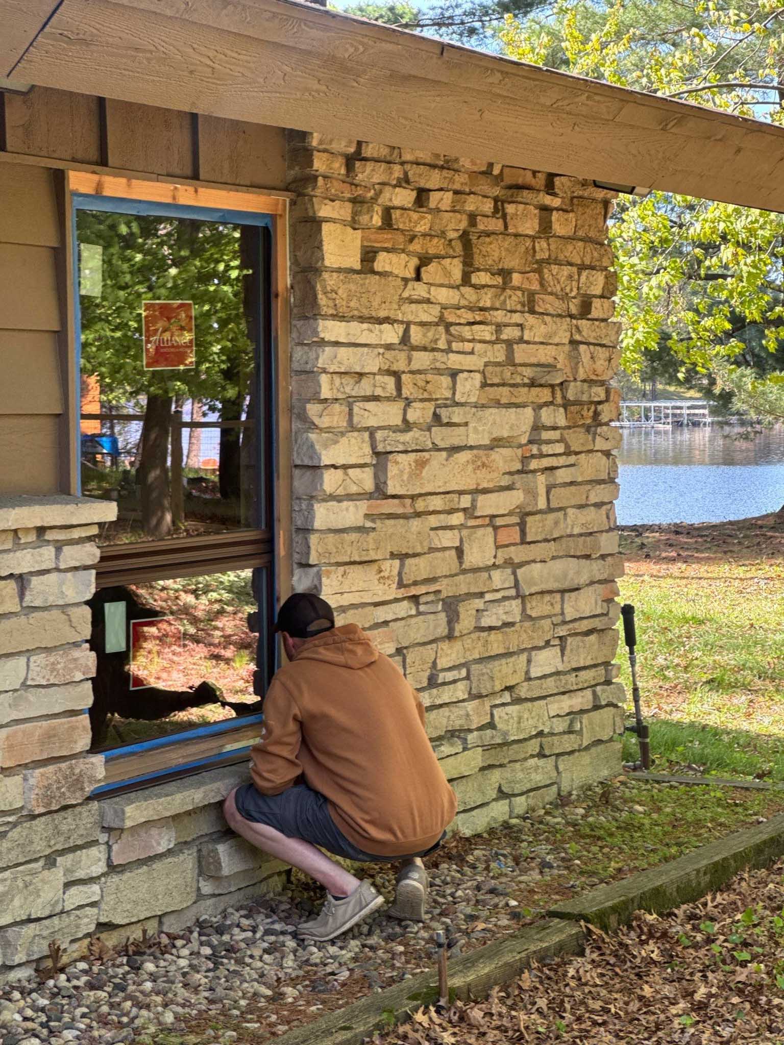 Person in a brown hoodie crouches by a stone building’s Replacement Windows Wisconsin near a lake.