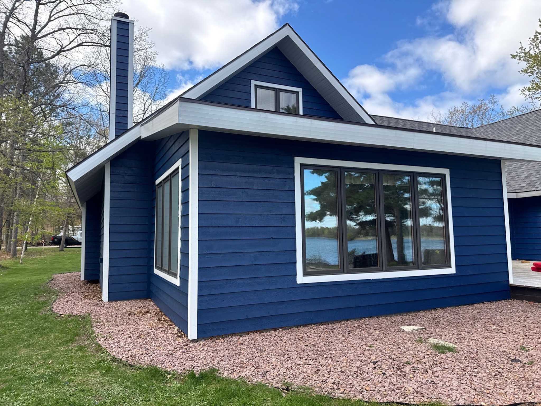 Blue house with large Replacement Windows Wisconsin, trees, rocks, and a lake in the background.