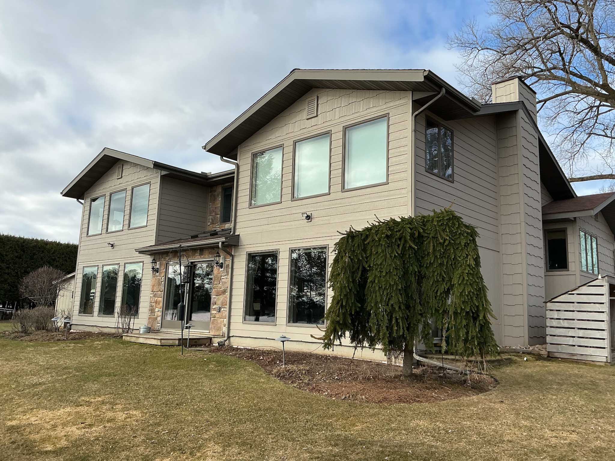 Two-story house with beige siding, replacement windows, and trimmed lawn in Wisconsin under clouds.